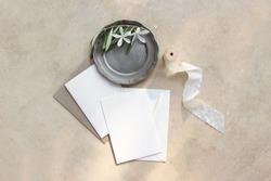 Summer wedding stationery mock-up scene. Blank greeting cards, envelopes, silver plate with olive branch and white flowers and silk ribbon. Marble background in sunlight, shadows. Flat lay, top view.