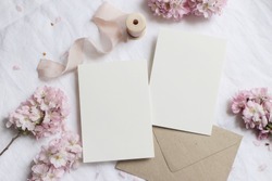 Wedding stationery mock-up scene. Blank greeting cards, envelope on linen tablecloth background with pink blossoming cherry tree branches and ribbon. Feminine still life composition. Flat lay,top view
