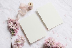 Wedding spring styled stock photo. Feminine desktop mockup scene with pink blossoming Japanese cherry tree branches and blank paper greeting cards on white linen table background. Flat lay, top view.