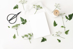 Wedding, birthday desktop mock-up scene. Blank greeting card, black vintage scissors, silk ribbon and cow parsley leaves and flowers. White table background. Flat lay, top view. Feminine stationery 