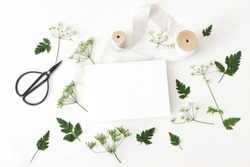 Wedding, birthday desktop stationery mock-up scene. Blank greeting card, black vintage scissors, silk ribbon and cow parsley leaves, flowers. White table background. Horizontal flat lay, top view. 