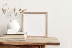 Square wooden frame mockup on vintage bench, table. Modern white ceramic vase with dry grass, books and busines card. White wall background. Scandinavian interior.