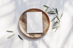 Summer wedding stationery mock-up scene. Blank greeting card, wooden plate, olive tree leaves and branches in sunlight. White table  background with palm shadows. Feminine flat lay, top view.