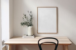 Home office concept. Old books, empty vertical wooden picture frame mockup hanging on white wall. Wooden desk, table. Vase with olive branches. Elegant working space. Scandinavian interior design.