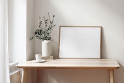Home office concept. Empty horizontal wooden picture frame mockup. Cup of coffee on wooden table. White wall background. Vase with olive branches. Elegant working space. Scandinavian interior design.
