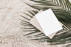 Summer stationery still life. Closeup of blank card mock-up and craft envelope on green date palm leaves. Sandy beach background or desert. Flat lay, top view. Tropical vacation concept. 