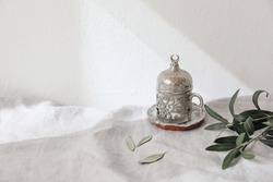 Decorative silver Turkish cup of tea. Green olive tree branches on white linen table cloth in sunlight. Festive still life for Muslim community holy month Ramadan Kareem. Blurred background, shadows.