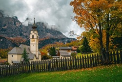 Majestic autumn scenery with colorful larch forest on the cliffs. Charming alpine village with beautiful church. Great hiking and touristic places, Colfosco, Dolomites, Italy, Europe
