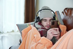 Young man warmly dressed is feeling cold, covered with warm blanket, sitting on the couch at cold home. Have problem with health, central heating