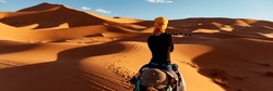 A young women in a yellow cap rides a camel  through the dunes in the Sahara Desert. View of the woman from behind, in the background, small silhouettes of other tourists. Merzouga, Morocco