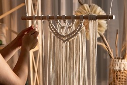 Woman craftsman weaves macrame from light cotton threads. Women's hands knit a beautiful lace ornament from knots in home workshop. Creative process handcraft, textile crafts. Close up.