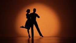 Couple of dancers approach each other and begin to dance Argentine tango. Elements of latin ballroom dance in studio with orange brown background. Dark silhouettes. Slow motion ready, 4K at 59.94fps.