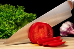 A metal knife slicing a red ripe tomato on a wooden board. Close up of a sharp knife blade cutting tomato on slices, green salad and garlic on a black isolated background.