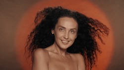 Seminude woman on an orange background with a circular light. Thick, long curly hair of a woman fluttering in the wind. The concept of beauty, hair styling, care.