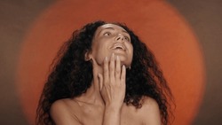 A woman enjoys the softness and silkiness of her skin, touches her neck. Portrait of a seminude curly woman in the studio on an orange background with circular light.