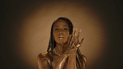 A woman with golden lips, golden skin and golden chains in her hair in the studio on a brown background with circular light. Arms of a seminude African American woman in liquid gold, or gold paint.