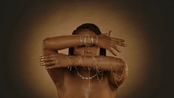 An African American woman demonstrates her hands with gold chain bracelets, her fingertips and nails are painted with gold paint. Seminude woman in the studio on a brown background with circular light