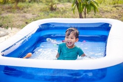 Portrait of asian child, kid or little boy to smile, bath, play water or funny activity in big blue portable inflatable swimming pool at outdoor, backyard or garden in summer evening. Look fun, happy.