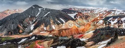 Panoramic amazing Icelandic landscape of colorful rainbow volcanic Landmannalaugar mountains, at famous Laugavegur hiking trail with dramatic snowy sky, and red volcano soil in Iceland.
