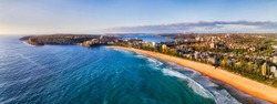 Long strip of clean yellow sandy beach of Manly - part of Sydney famous Northern Beaches seen from mid-air with distant Sydney harbour and Manly waterfront.