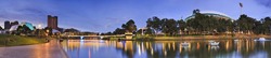 panorama of Adelaide city centre over Torrens river at sunrise. From CBD high-rises to oval in public park with blurred water and reflecting lights.