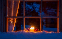 Snowy evening window of a wooden house. On the windowsill are Christmas decorations and burning candles
