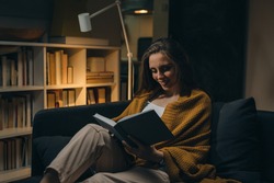 woman relaxing on sofa and reading book. evening moody ambience. she is enjoying time during weekend