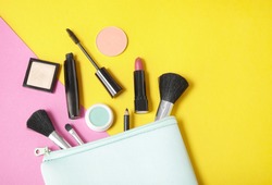 Make up products spilling out of a pastel blue cosmetics bag, on to a bright yellow and pink background with empty space at side