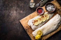 Raw cod fish fillet with lemon slices and herbal salt on rustic wooden cutting board on dark background, top view. Fish cooking preparation. Healthy diet food. Border. Copy space