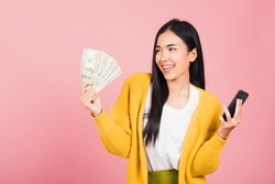Portrait Asian happy beautiful young woman teen shopper smiling standing excited holding online smart mobile phone and dollar money banknotes on hand in summer, studio shot isolated on pink background