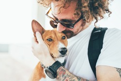 Young trendy hipster with tattoos crazy curly hair with his best friend a cute small basenji puppy gives him a tender kiss on ears while hugs him with his arms