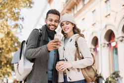 Beautiful happy couple using smartphone. Young joyful smiling woman and man looking at mobile phone in a city in autumn.  Technology, travel, tourism, students concept