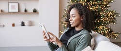 Young woman on sofa using smartphone, relaxing at home during Christmas holidays. Young african american woman looking at mobile phone. Technology, connection, communication, modern lifestyle concepts