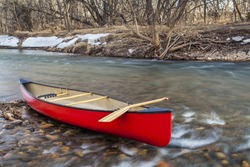 red canoe with a wooden paddle on river shore in winter or early spring - Cache la Poudre River, Fort Collins, Colorado