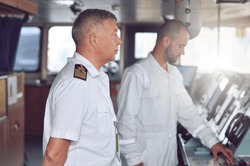 The captain of a cargo ship in a white shirt and shoulder straps on the bridge gives instructions to the navigator