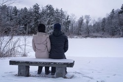 A couple sitting on a snow covered bench overlooking a frozen pond within the white memorial conservation area in Litchfield Connecticut on a cold snowing winter day in new england.