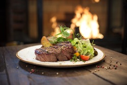 Food - Beef dinner - Delicious grilled stake and potatoes served on a wooden table, fireplace on background. Big steak meat dish on a main course plate