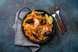 Classic dish of Spain, seafood paella in traditional pan on rustic blue concrete background top view. Spanish paella with shrimps, clamps, mussels, green peas and fresh lemon wedges from above 
