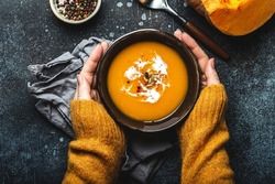 Female hands in yellow knitted sweater holding a bowl with pumpkin cream soup on dark stone background with spoon decorated with cut fresh pumpkin, top view. Autumn cozy dinner concept 