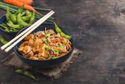Asian noodles with chicken, vegetables in bowl, rustic wooden background. Space for text. Soba noodles, teriyaki chicken, edamame, chopsticks. Closeup. Asian style dinner. Chinese/Japanese noodles