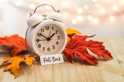 Fall Back Daylight Saving Time concept with white clock and autumn leaves, soft bokeh background on wooden board