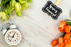 Spring Forward Daylight Saving Time concept with text on marble flat lay.  White alarm clock with tulips.