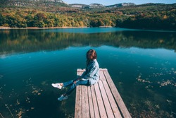 Young woman sitting on peer and enjoying lake view in autumn. Good sunny day for resting outdoors and travel.