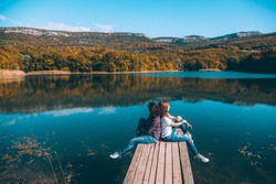 Two friends sitting on peer and enjoying lake view in autumn. Good sunny day for resting outdoors and travel.