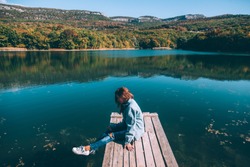 Young woman sitting on peer and enjoying lake view in autumn. Good sunny day for resting outdoors and travel.