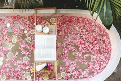 Top view of bath tub with flower petals and lemon slices. Book, candles and beauty product on a tray. Organic spa relaxation in luxury Bali outdoor bathroom.