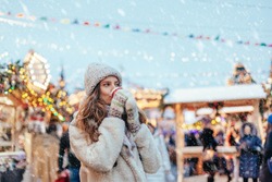 Girl drinking hot coffee while walking in Christmas market decorated with holiday lights in the evening. Feeling happy in big city. Spending winter vacations in Red square, Moscow, Russia.
