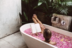 Woman reading book while relaxing in bath tub with flower petals. Organic spa relaxation in luxury Bali outdoor bath.