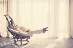 Young woman at home sitting on modern chair in front of window relaxing in her living room reading book, instagram toning