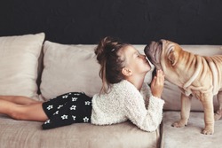 Home portrait of cute child kissing puppy of Chinese Shar Pei dog on the sofa against black wall